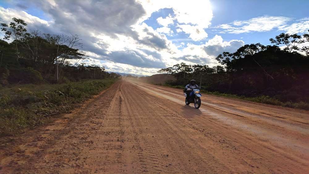 Lone motorcyclist on a wide red dirt road through low forest and big sky