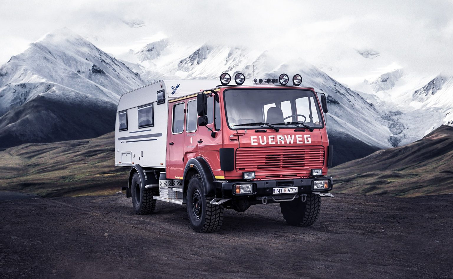 Red expedition truck with white cabin on dark ground in front of snowy mountains