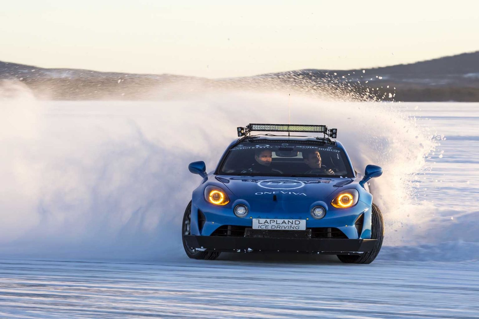 Blue Alpine sports car drifting on ice, spraying snow, hills in the distance