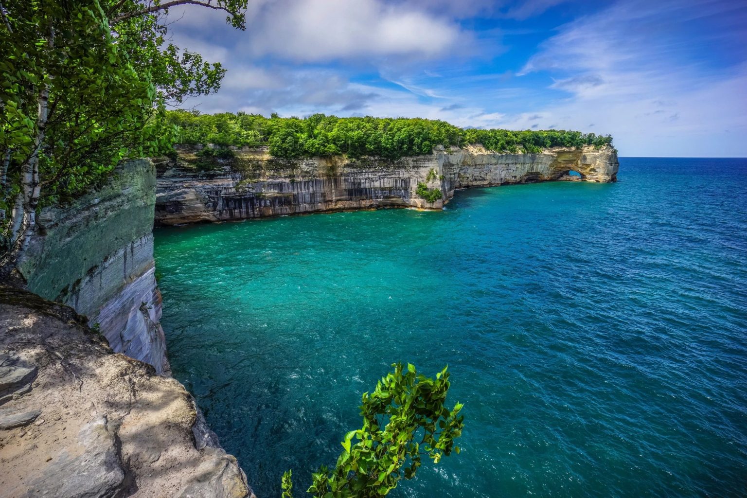 Turquoise lake water beside layered sandstone cliffs topped with forest, Pictured Rocks on Lake Superior