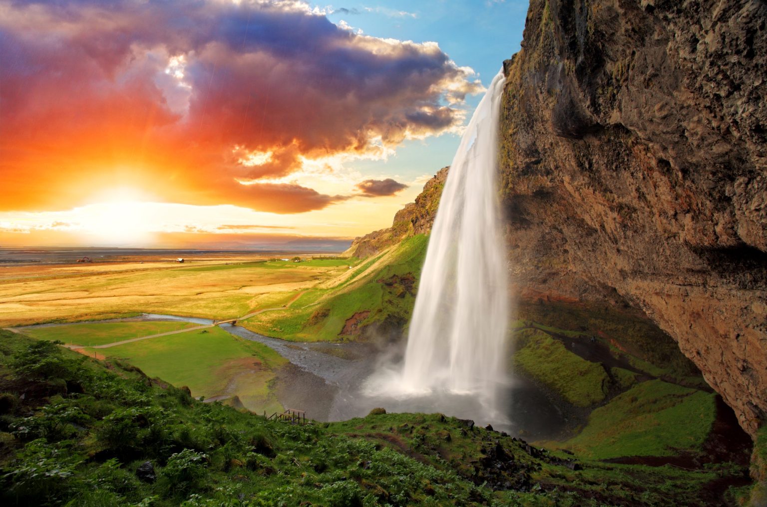 Slender waterfall beside a cliff at sunset over green Icelandic farmland