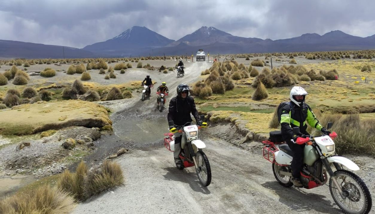 Motorcyclists riding a muddy track through highland grass towards a volcano range