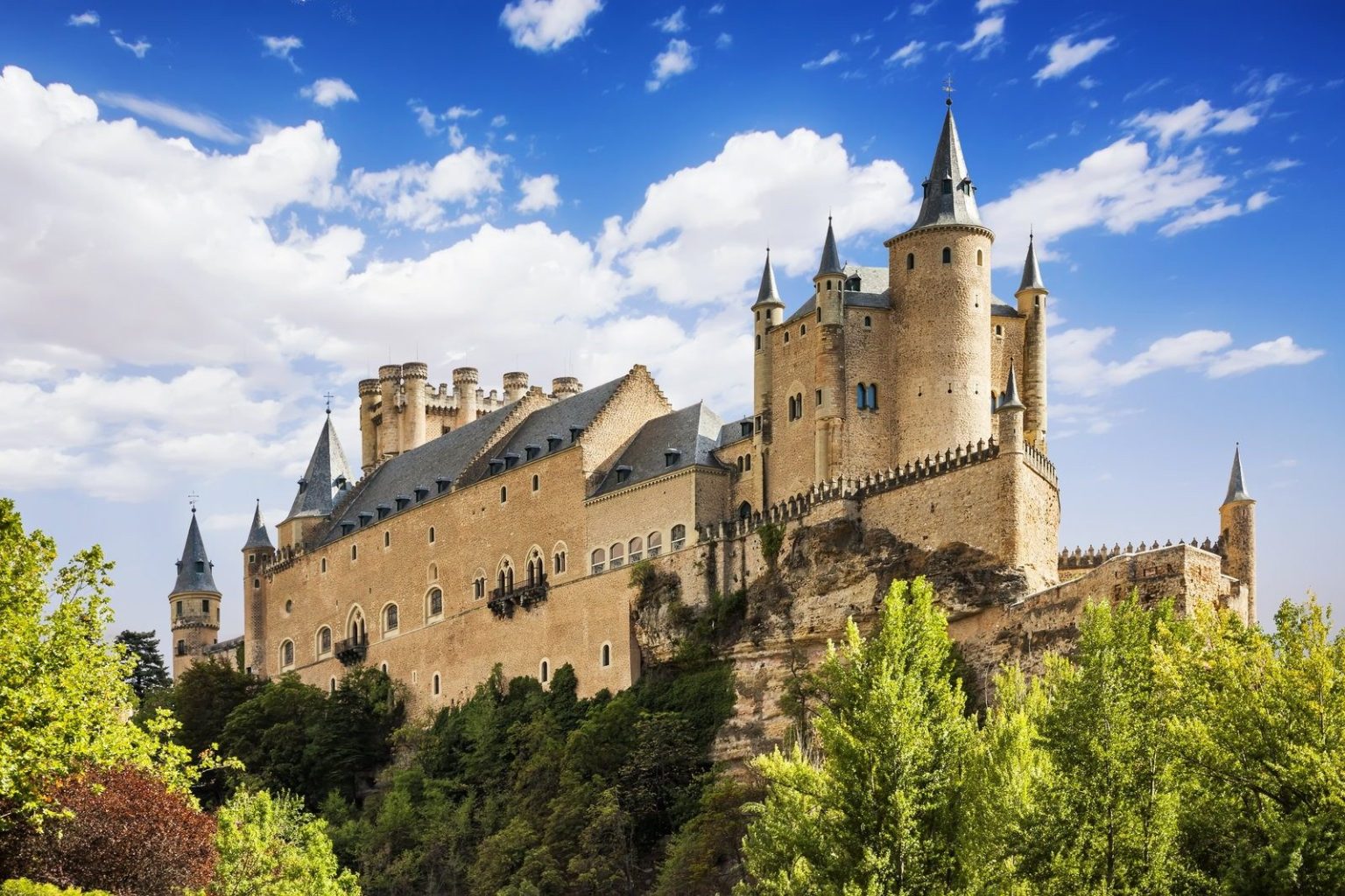 Hilltop castle with slate turrets and towers above trees under a blue sky, Segovia