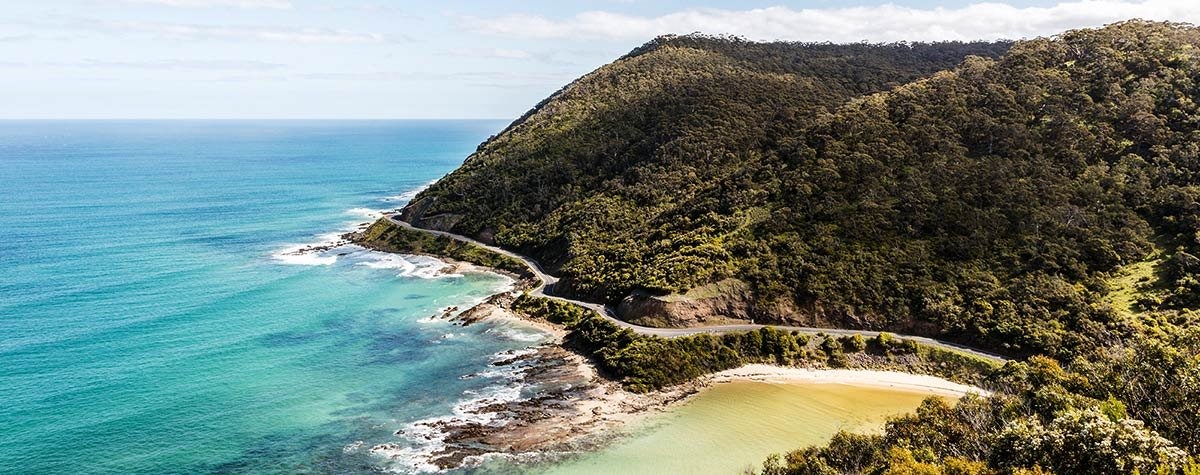 Coastal road winding along a forested headland above turquoise sea and a sandy cove