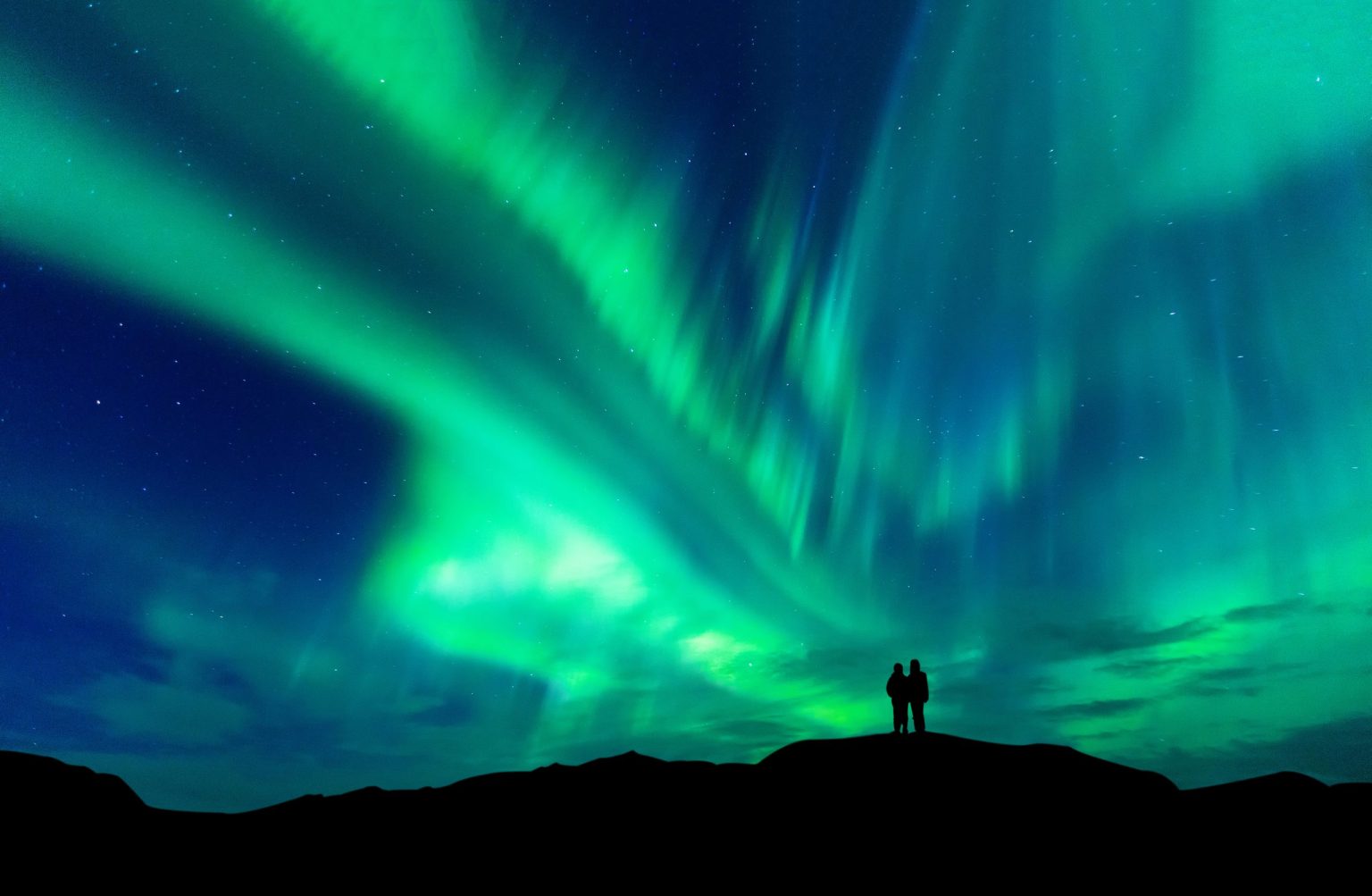 Two people silhouetted on a hilltop beneath bright green northern lights