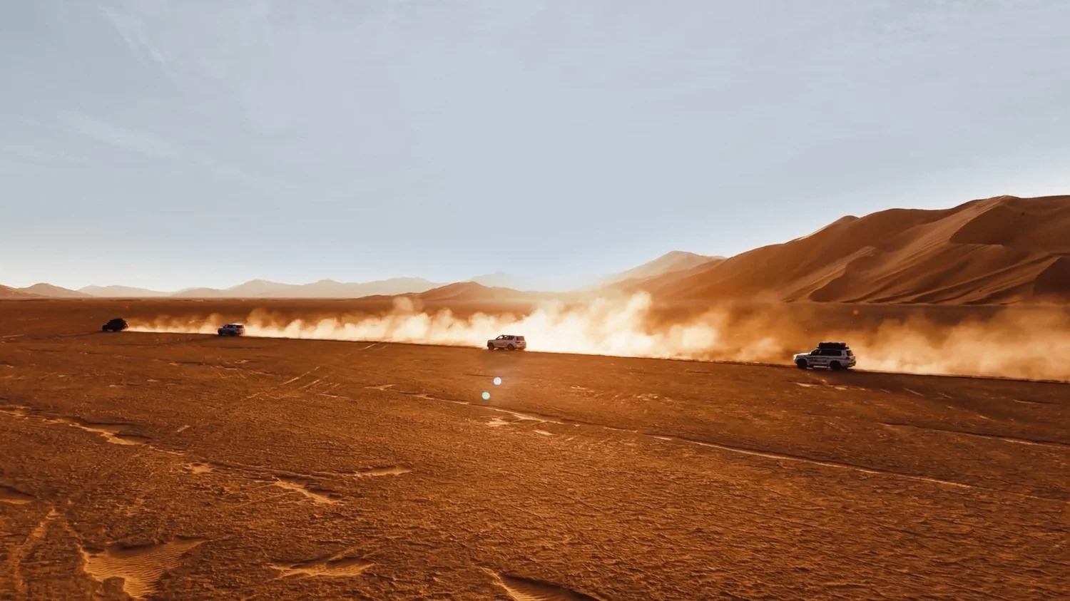 Convoy of 4WD vehicles kicking up dust across a flat desert plain by tall dunes