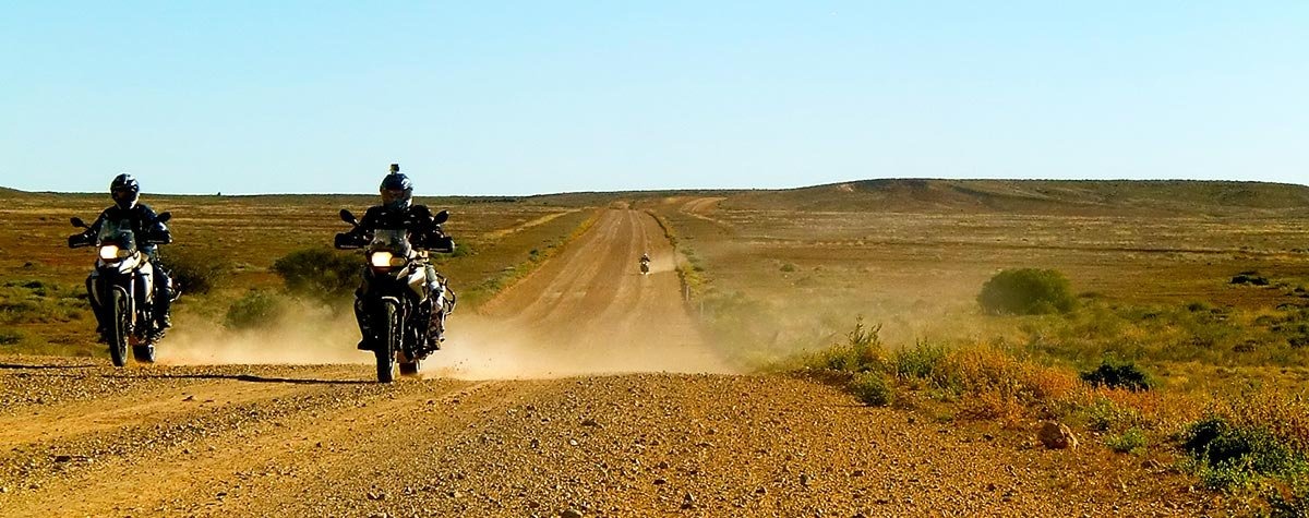 Two motorcyclists riding side by side on a dusty outback gravel road