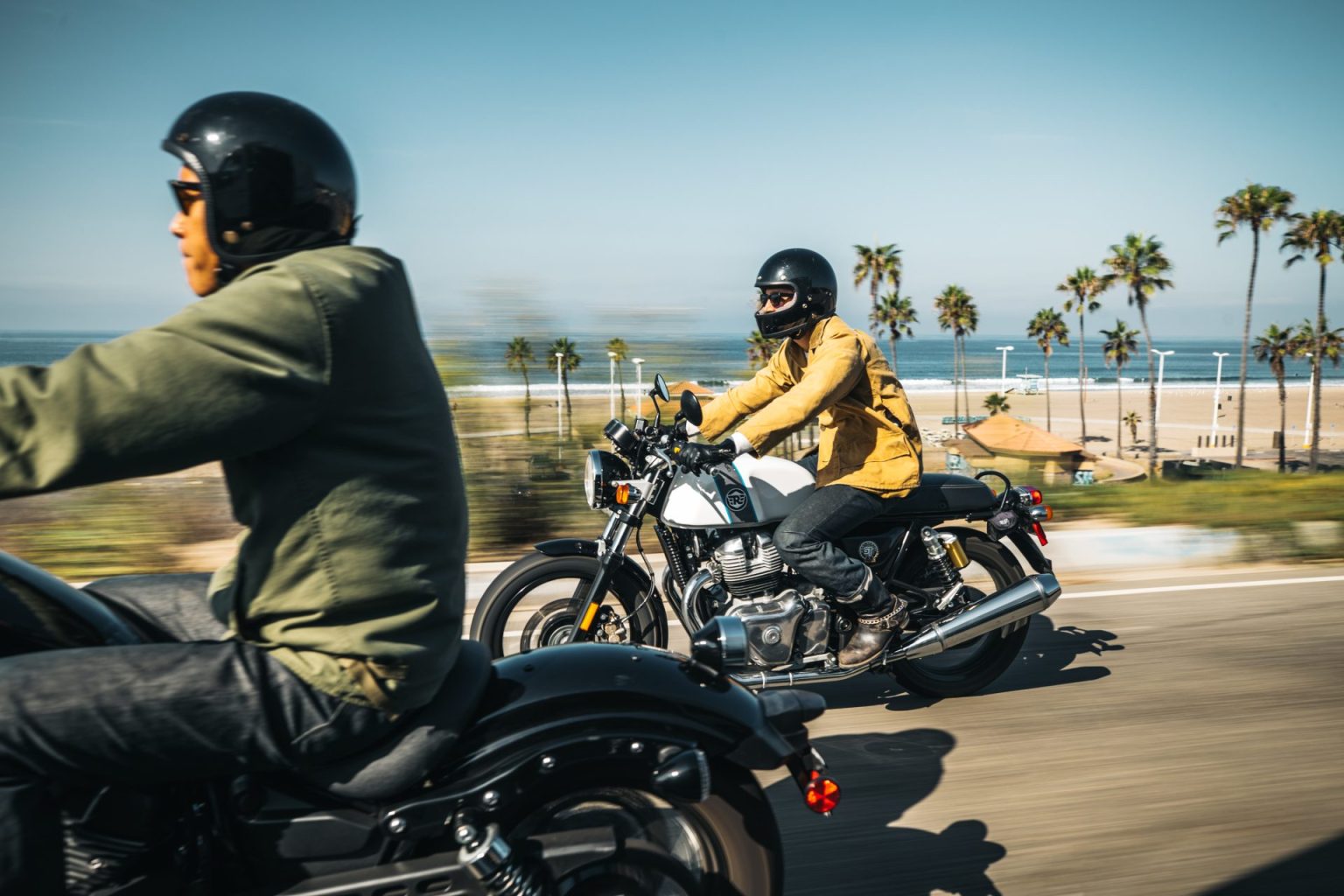 Two riders on motorcycles passing palm trees and a beach in Southern California