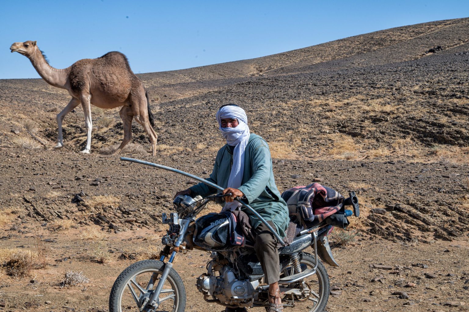 Local in a blue turban with a camel beside a dirt bike in the Sahara