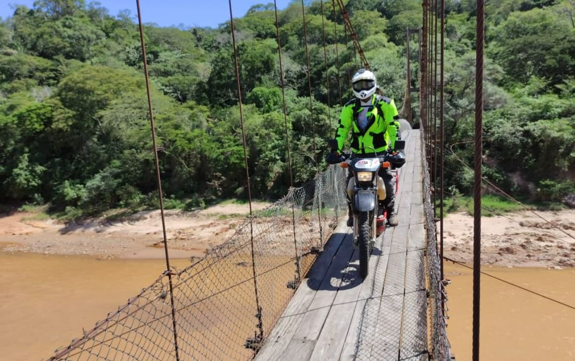 Motorcyclist riding across a narrow wooden suspension bridge above a muddy river