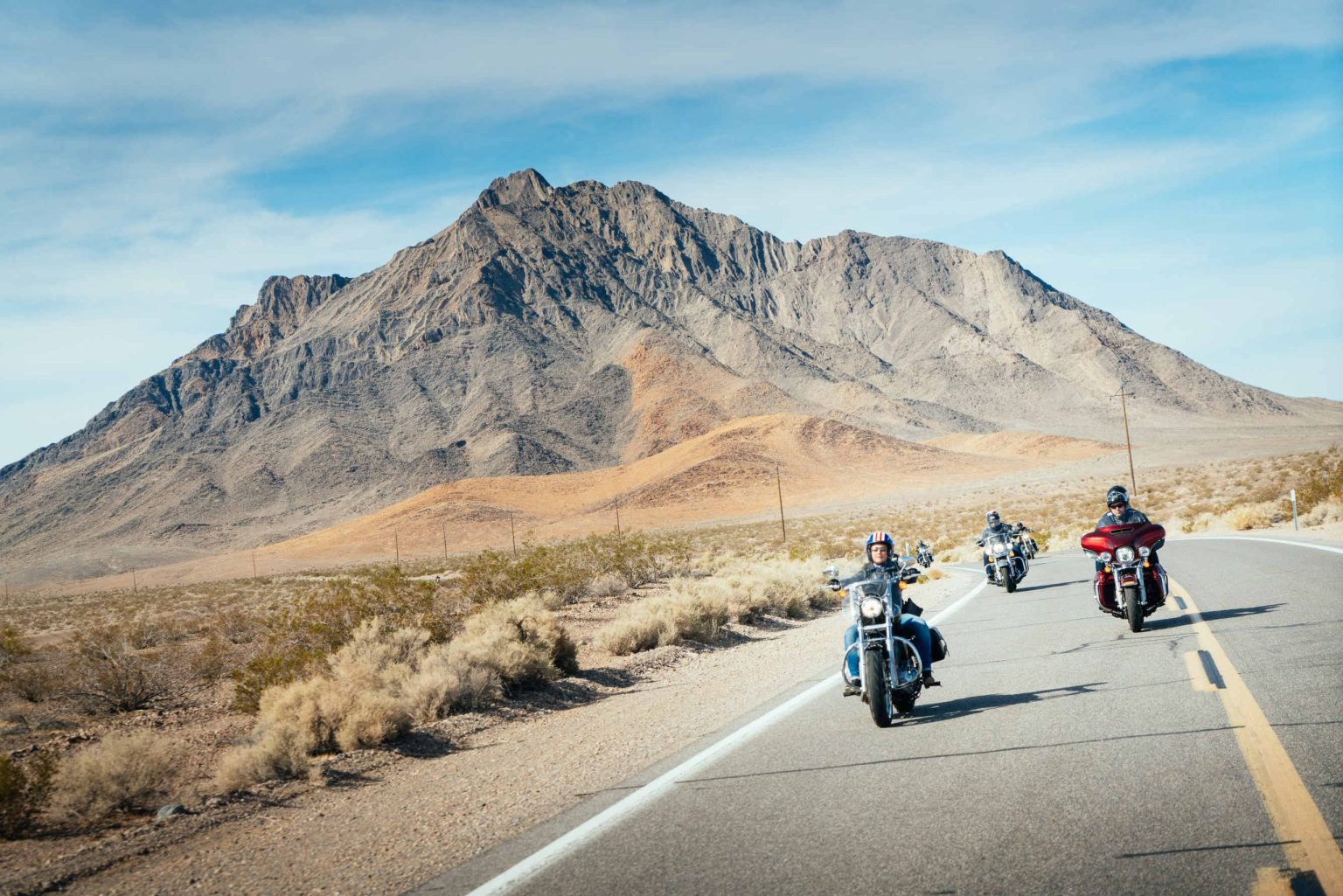Motorcyclists riding a desert highway toward a rugged mountain peak